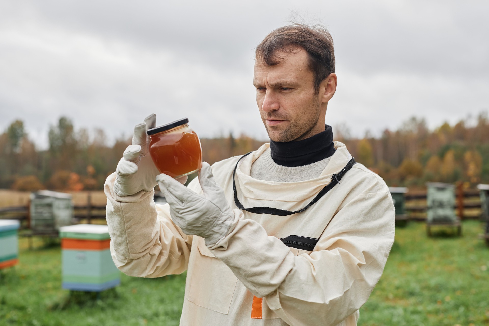 Male Beekeeper in Suit Holding Honey Jar against Grey Sky at Apiary Farm